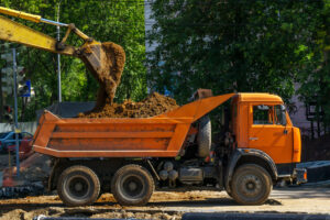 A backhoe dumps soil into the back of an orange dump truck