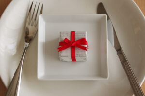 A small square gift with a red bow on a square white plate with fork and knife next to the plate.