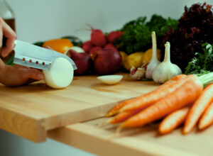 A knife chopping an onion on a wooden board with carrots, garlic and other vegetables around