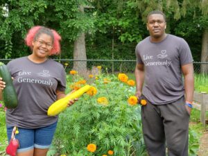 Two young people standing in front of a garden, one holding a yellow squash and a green squash