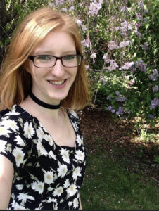 A young woman with red hair and glasses, wearing a black dress with white flowers