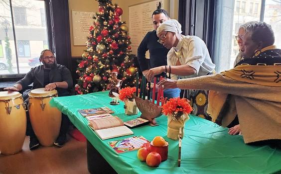 Four people standing behind a table with a green table cloth and Kwanzaa symbols. Two people are lighting candles on a kinara.